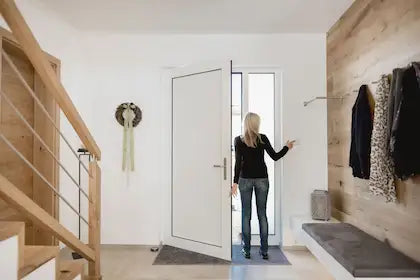 Een vrouw bedient een witte Loxone Touch Pure schakelaar bij een openstaande voordeur in een modern interieur om alle technieken uit te schakelen bij vertrek, begeleid door Kompanjonn.
