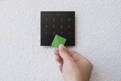 Een hand houdt een groene Loxone NFC-key tegen een zwart NFC Code Touch bedieningspaneel op een witte buitenmuur voor veilige en slimme toegangscontrole, geadviseerd door Kompanjonn.