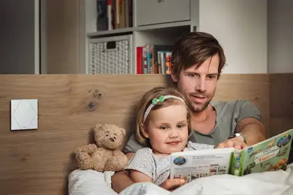 Sfeervolle slaapkamer met een vader en dochter die lezen, waarbij een witte Loxone Touch Pure schakelaar op het houten hoofdbord zorgt voor eenvoudige bediening van de slimme verlichting via Kompanjonn.